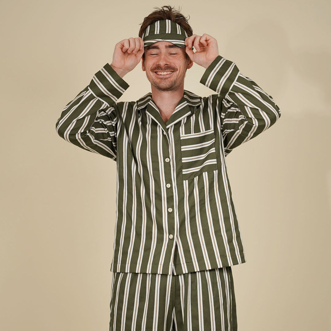 A man wearing a matching green and white vertically striped pajama set with a headband, posing with his hands on the headband against a solid beige background.