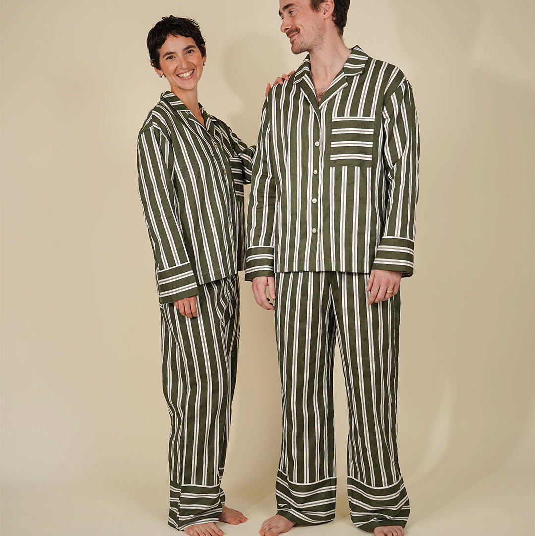 A man and a woman wearing matching striped pajama sets, standing barefoot against a plain beige studio background. The pajamas feature vertical stripes in dark green and off-white.
